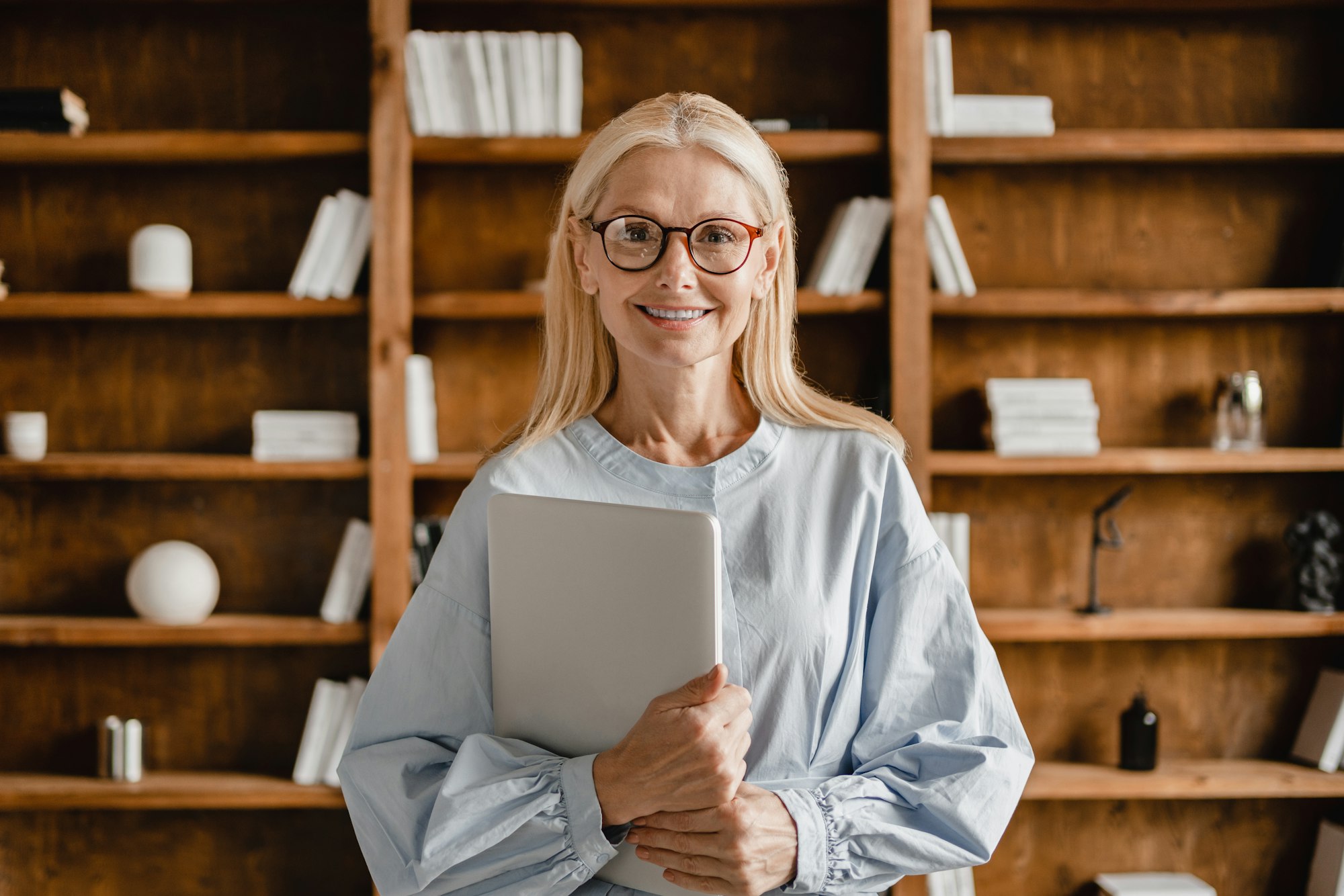 Businesswoman teacher boss freelancer holding laptop looking at camera in glasses in office library